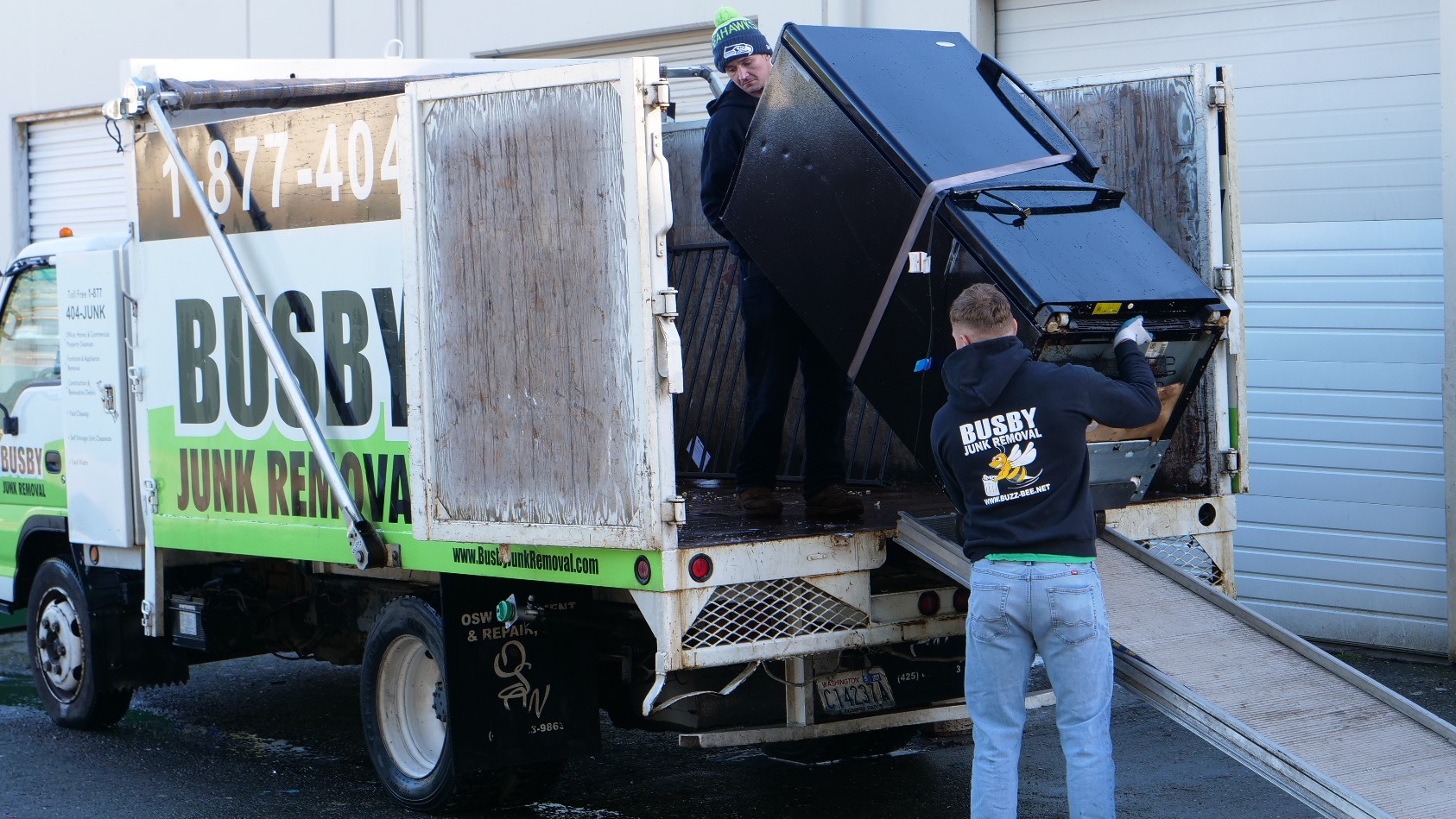 Professional team from Busby Junk Removal efficiently removing an old refrigerator from a residential home. The image shows two uniformed workers, wearing safety gloves and boots, carefully maneuvering the large appliance through a spacious, well-lit kitchen. The refrigerator is being carried with the aid of a sturdy dolly, ensuring safe and damage-free removal. In the background, there are clean, empty countertops and a glimpse of the open front door, indicating the pathway for removal. The scene conveys the company's commitment to professionalism, safety, and customer satisfaction in household appliance removal services